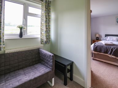A small sitting area with a gray sofa and a black table near a window with patterned curtains overlooking a green field and a bedroom with a bed and bedside lamp at Aberdauddwr in Lampeter