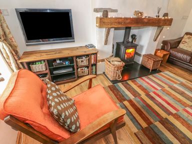 A living room with a wall-mounted TV wooden cabinet wicker baskets a wood-burning stove and an orange armchair at Rosemary Cottage in Clawthorpe near Burton-in-Kendal