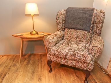 A floral armchair with a gray blanket next to a wooden side table with a lamp at Rosemary Cottage in Clawthorpe near Burton-in-Kendal