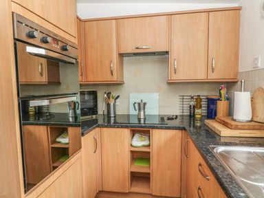 A kitchen with wooden cabinets a black countertop an oven a microwave cooking utensils and a stainless steel sink at Rosemary Cottage in Clawthorpe near Burton-in-Kendal