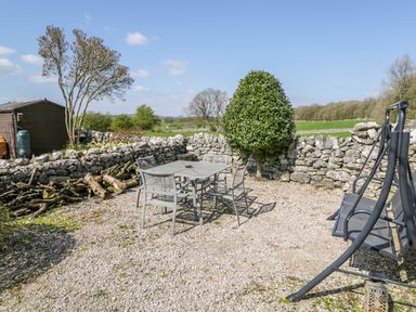An outdoor patio area with a metal table and chairs near a stone wall with logs and a swing seat at Rosemary Cottage in Clawthorpe near Burton-in-Kendal