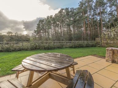 A wooden round table with attached benches on a tiled patio overlooking a grassy yard and trees at Mill House in Lowick