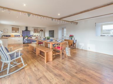 A spacious dining room and kitchen with wooden floor a dining table with benches and chairs a rocking chair and kitchen cabinets at Mill House in Lowick