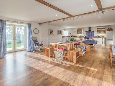 A dining area with a wooden table and benches set with plates and glasses next to a kitchen with cupboards a stove a fridge and a microwave at Mill House in Lowick