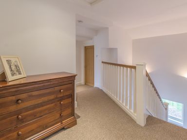 An upstairs hallway with a wooden chest of drawers a framed picture on top a white banister and a closed wooden door at Mill House in Lowick