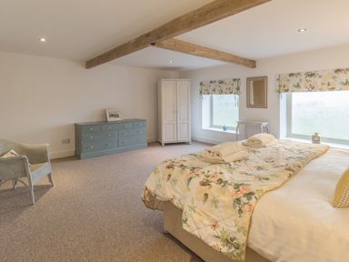 A bedroom with a bed covered with floral bedding two windows with floral blinds a chest of drawers a white wardrobe and a wicker chair at Mill House in Lowick