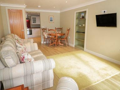 A living room with patterned sofas a wooden dining table with chairs a wall-mounted TV and a kitchen visible through a doorway at Bryn Y Don Cottage in Benllech