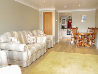 A living room with a striped sofa, floral cushions, wooden dining table with chairs and kitchen in the background at Bryn Y Don Cottage in Benllech