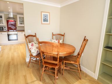 A dining area with a round wooden table and four wooden chairs with cushions next to a kitchen with white cabinets at Bryn Y Don Cottage in Benllech
