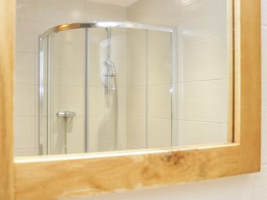 A bathroom shower enclosure with glass doors and chrome fixtures reflected in a wooden framed mirror at Bryn Y Don Cottage in Benllech