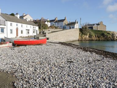 A pebble beach with a red boat on the shore and white houses on a street by the water at Bryn Y Don Cottage in Benllech