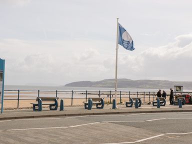 Benches along a beachfront promenade with a flagpole flying a blue flag and people near the railing at Bryn Y Don Cottage in Benllech