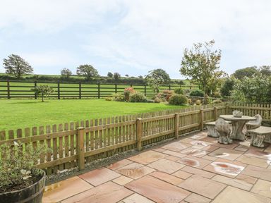 A patio with stone table and benches overlooking a fenced grassy garden and trees at Ty Ni in Caeathro near Caernarfon