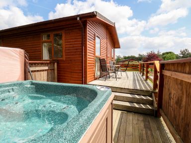 A wooden deck with a hot tub and a table with chairs outside a wooden cabin at Callow Lodge 5 in Beaconsfield Holiday Park