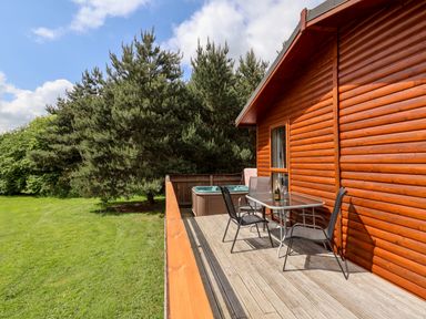 A wooden deck with a glass table and two chairs next to a wooden cabin with a hot tub and green trees at Callow Lodge 5 in Beaconsfield Holiday Park