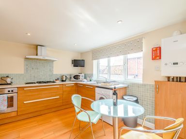 A kitchen with wooden cabinets a glass table two chairs a washing machine microwave and a window at Mermaid Cottage in Whitby