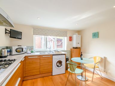 A kitchen with wooden cabinets a washing machine a round glass table with two chairs and a window at Mermaid Cottage in Whitby