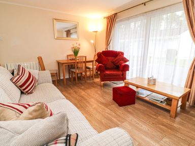 A living room with a sofa and armchair near a wooden table with chairs and a large window with curtains at Peacehaven Rosecraddoc Lodge near Liskeard
