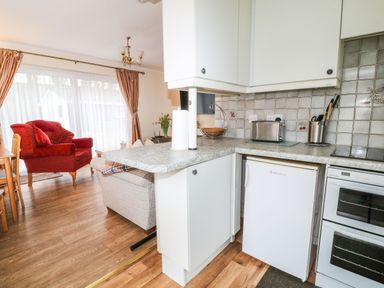 A kitchen counter with white cabinets and appliances next to a living area with a red armchair and wooden floor at Peacehaven Rosecraddoc Lodge near Liskeard