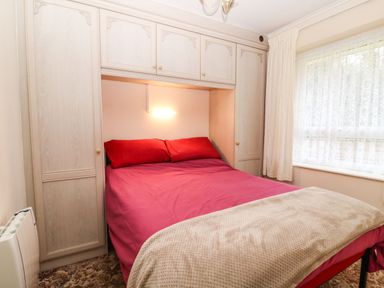 A bedroom with a bed with red bedding and pillows built into a wooden wardrobe with a window covered by curtains at Peacehaven Rosecraddoc Lodge near Liskeard