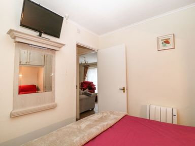 A bedroom with a bed covered in a red blanket a wall-mounted television and an open door showing a living room with sofas at Peacehaven in Rosecraddoc Lodge near Liskeard