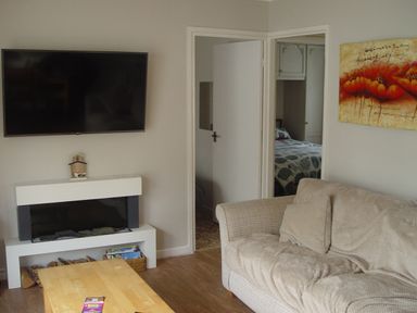 A living room with a mounted TV above a fireplace a beige couch with cushions a wooden coffee table and an open door showing a bedroom at Peacehaven Rosecraddoc Lodge near Liskeard