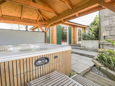 An outdoor covered hot tub with wooden paneling next to a patio and a wooden building with green shutters at Eden Lodge in Bardsea