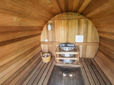 An interior of a wooden barrel sauna with benches a heater a bucket and a thermometer at Eden Lodge in Bardsea