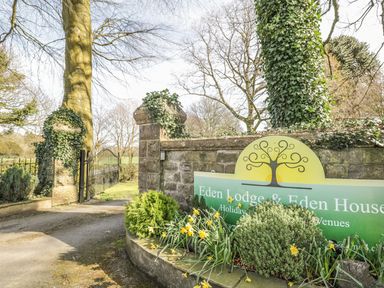 Entrance gate with stone pillars covered in ivy and a sign for Eden Lodge and Eden House surrounded by plants and daffodils at Eden Lodge in Bardsea