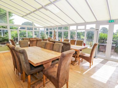 A dining room with two wooden tables surrounded by upholstered chairs in a room with large windows at Eden Lodge in Bardsea