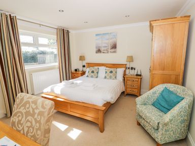 A bedroom with a wooden double bed with two bedside tables lamps and an armoire next to a window with striped curtains and a patterned armchair at Eden Lodge in Bardsea