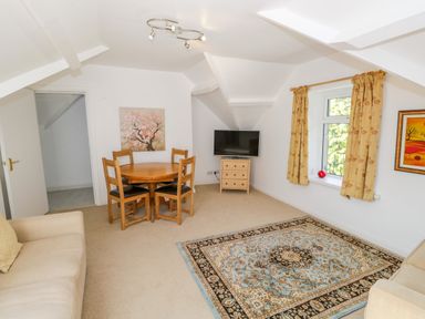 A living room with a round wooden table and chairs a television on a small chest of drawers a patterned rug and beige sofas at Eden Lodge in Bardsea
