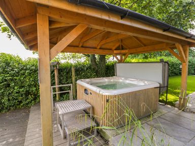 An outdoor hot tub under a wooden gazebo with steps beside it and greenery surrounding the area at Eden Lodge in Bardsea