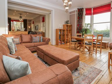 A living room with a brown sectional sofa and ottoman a wooden dining table with chairs and a wooden bookshelf by large windows with red blinds at Eden Lodge in Bardsea