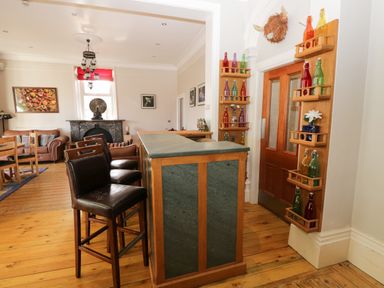 A room with a bar counter and three bar stools next to a seating area with sofas and a dining table at Eden Lodge in Bardsea