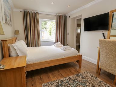 A bedroom with a wooden bed and bedside table a window with striped curtains a wall-mounted television a patterned chair and a rug on a wooden floor at Eden Lodge in Bardsea