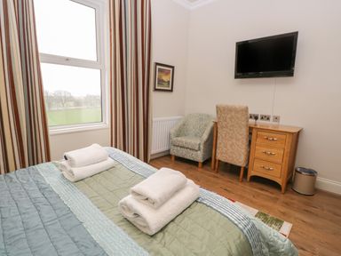A bedroom with a bed with folded towels a window with striped curtains a wooden desk with a chair an armchair and a wall-mounted TV at Eden Lodge in Bardsea