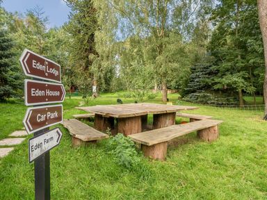 A wooden picnic table with benches on grass and signs pointing to Eden Lodge Eden House Car Park and Eden Farm in a wooded area at Eden Lodge Bardsea