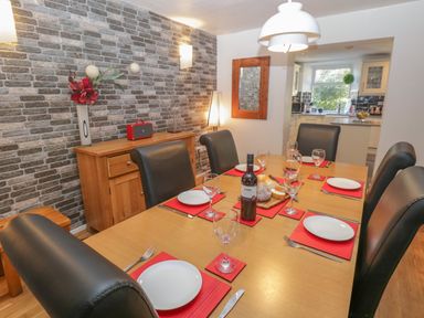 A dining room with a wooden table set with plates glasses and cutlery six black leather chairs a sideboard with a vase and a red radio and a kitchen visible in the background at 48 Oak Street Windermere