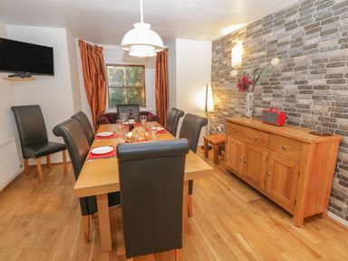 A dining room with a wooden table set with plates and glasses six black chairs a wooden sideboard with decorations and a wall-mounted television at 48 Oak Street in Windermere