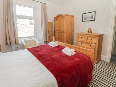 A bedroom with a double bed with white and red bedding a wooden wardrobe chest of drawers a white chair and a window with curtains at 48 Oak Street in Windermere