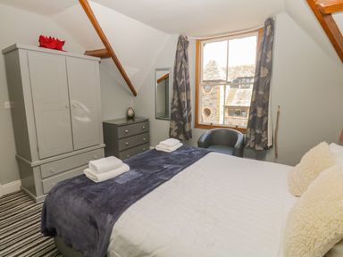A bedroom with a double bed, a gray wardrobe, a chest of drawers, a window with curtains, and a gray armchair at 48 Oak Street in Windermere
