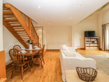A dining area with wooden table and chairs next to a staircase and a living room with white sofa and TV cabinet at Meader Barn in Peter Tavy near Tavistock