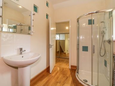 A bathroom with a pedestal sink, a mirror, a shower cubicle with glass doors and tiled walls, wooden floor and an open door showing a hallway at Meader Barn Peter Tavy near Tavistock