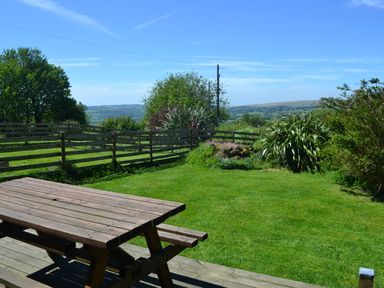 A wooden picnic table on a deck overlooking a fenced grassy garden with trees and hills in the background at Meader Barn in Peter Tavy near Tavistock