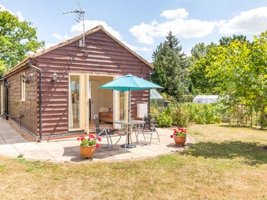 A small wooden house with a patio table, chairs, and umbrella in a garden at Norbank Garden Studio in Bressingham