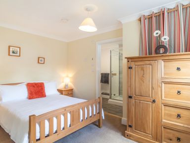 A bedroom with a wooden bed white bedding and orange cushion with wooden bedside table and lamp next to a wooden wardrobe with flowers and striped curtains at Norbank Garden Studio in Bressingham