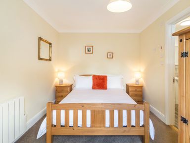 A bedroom with a wooden bed frame white bedding and an orange pillow two wooden bedside tables with lamps a wooden wardrobe a mirror and two small framed pictures on the wall at Norbank Garden Studio in Bressingham