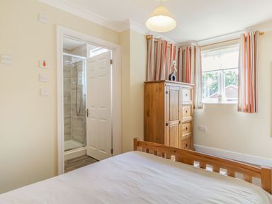 A bedroom with a wooden bed a wooden wardrobe striped curtains and an open door showing a shower at Norbank Garden Studio in Bressingham