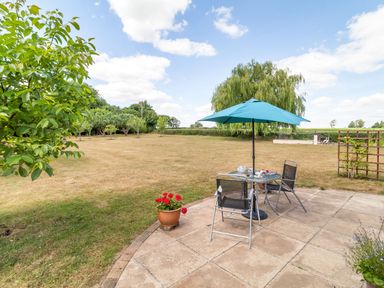 A garden patio with a table, chairs, and an umbrella overlooking a large grassy yard with trees at Norbank Garden Studio in Bressingham